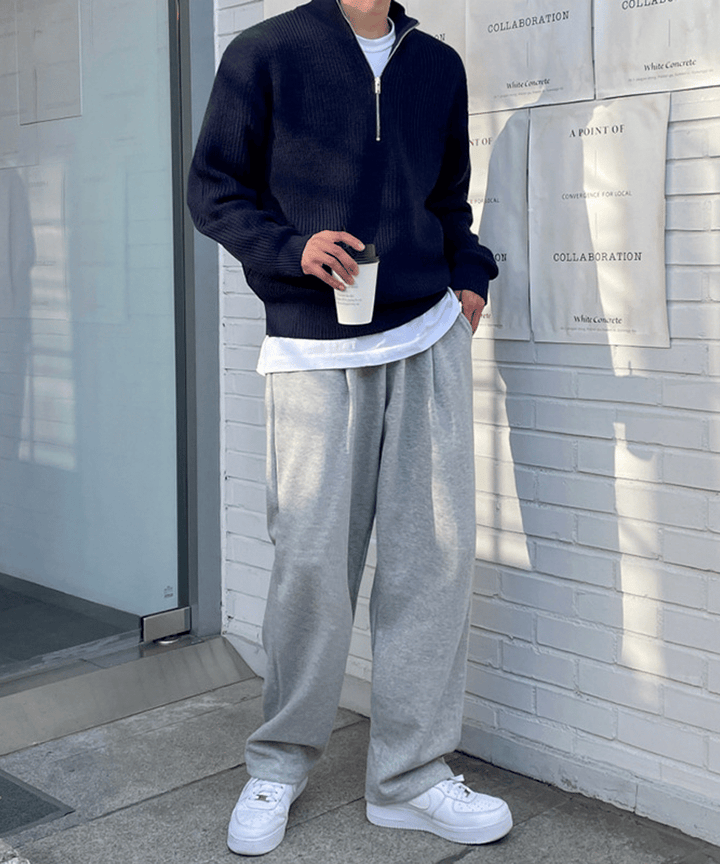 白いレンガ壁沿いに佇む男性のスタイリングカット。ネイビーのハーフジップセーターに、裾から覗く白Tシャツ、ライトグレーのルーズなワイドスウェットパンツ、足元にボリュームのある白スニーカーを合わせた都会的なカジュアルコーディネート。