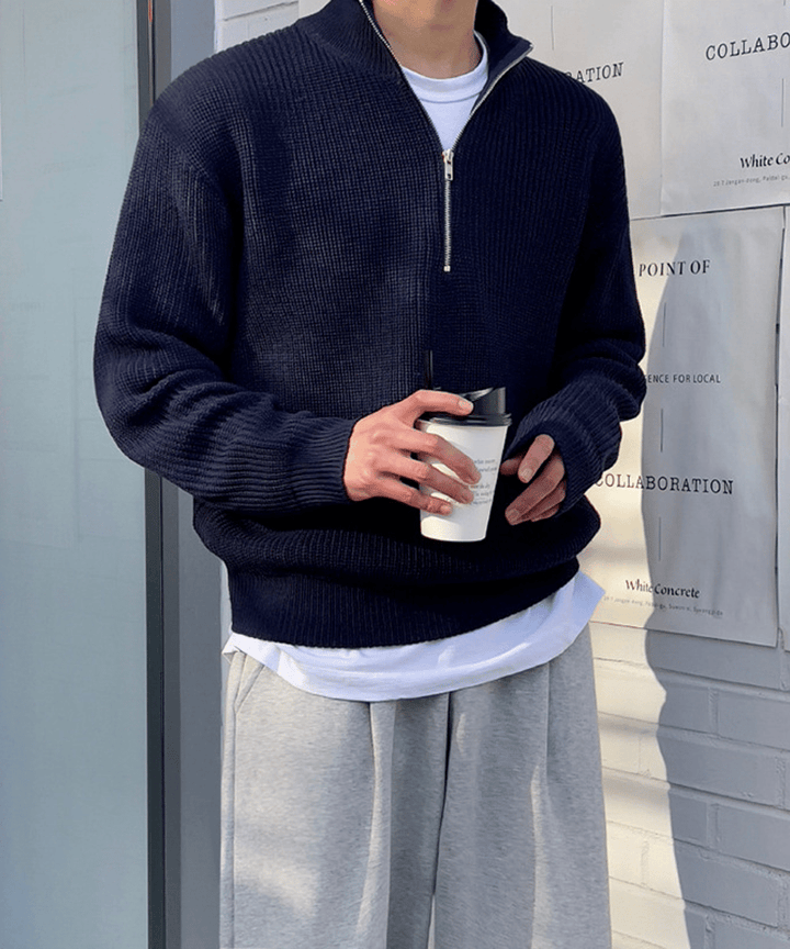 : ポスターが貼られた白いレンガ壁の前で、ネイビーのセーターを着用した男性のアップカット。首元のジッパーを少し開け、インナーの白Tシャツを覗かせながら、手元にテイクアウトのコーヒーカップを持ったリラックス感のあるポーズ。