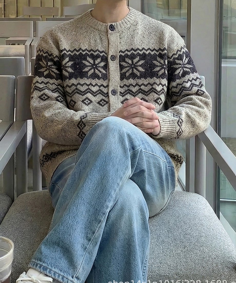 都会的なカフェの椅子に座り、両手を組んで足を組む男性のスタイリング。ベージュのノルディック柄カーディガンと淡色デニムを上品に合わせた、大人のリラックスカジュアルスタイル。
