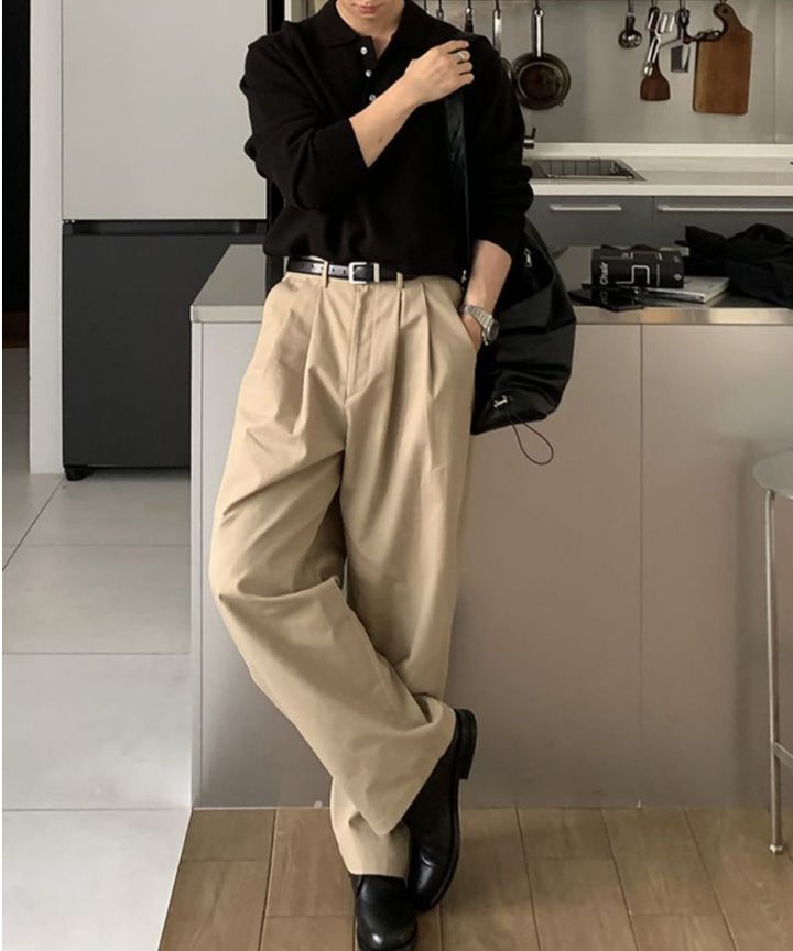のポロニットとベージュのタックワイドパンツに黒の革靴を合わせ、キッチンのカウンターに寄りかかって立つ人物の全身カット。