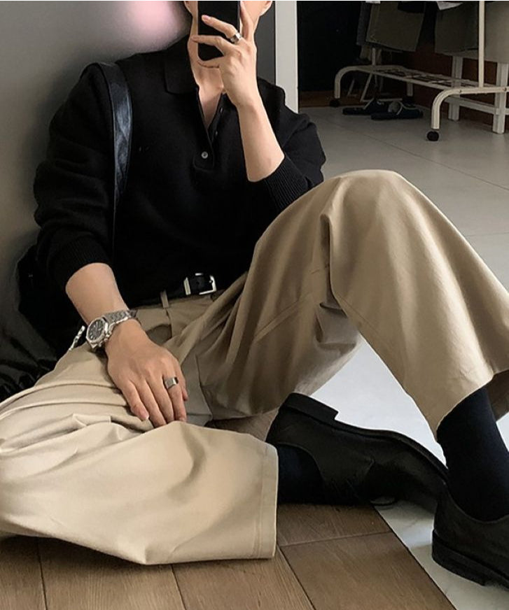 ブラックのポロニットにタック入りのベージュチノパンツ、足元に黒のレザーシューズとソックスを合わせ、床に座ってスマートフォンで自撮りをする人物の全身スタイリング。