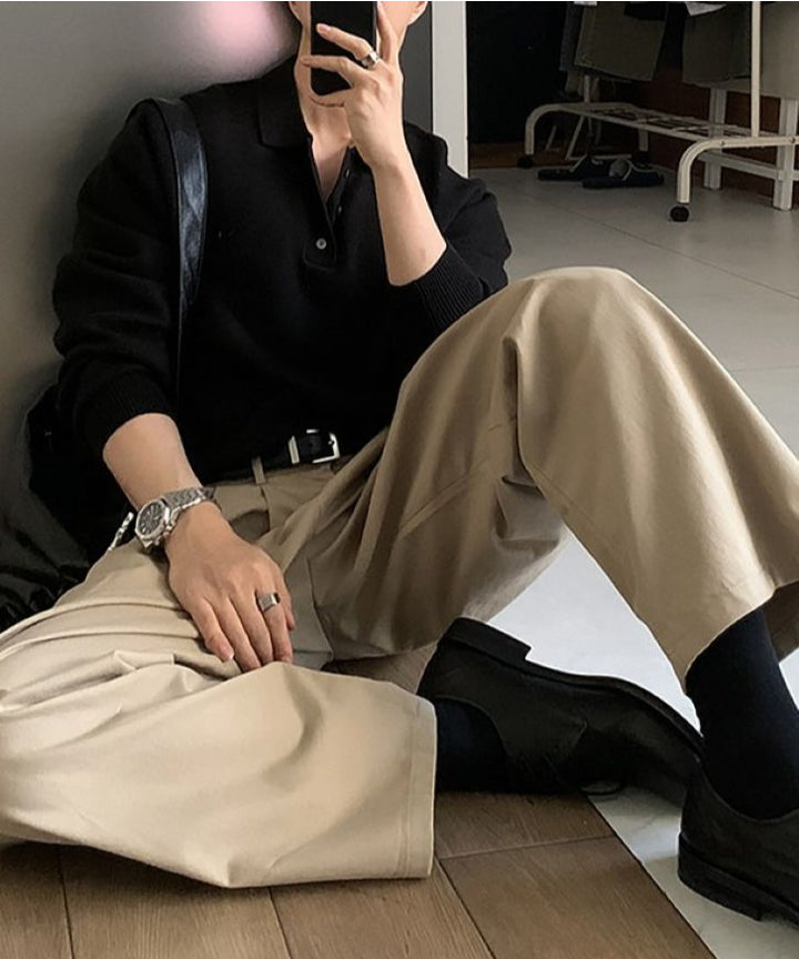 ブラックのポロニットにタック入りのベージュチノパンツ、足元に黒のレザーシューズとソックスを合わせ、床に座ってスマートフォンで自撮りをする人物の全身スタイリング。