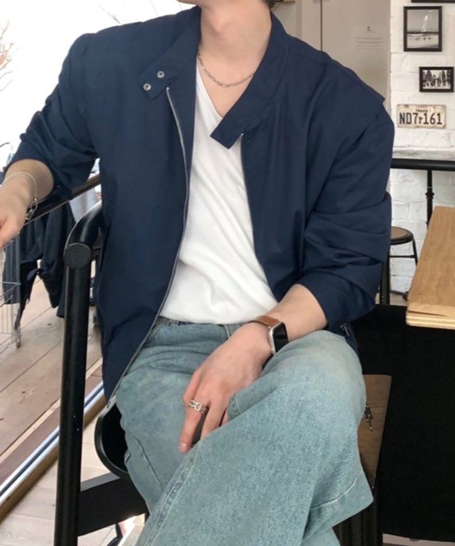 カフェのテラス席に腰掛け、足を組んでリラックスする男性のスタイリングカット。ネイビーのスタンドカラーブルゾンに白Tシャツ、淡色ワイドデニムを合わせた大人のアーバンカジュアルルック。