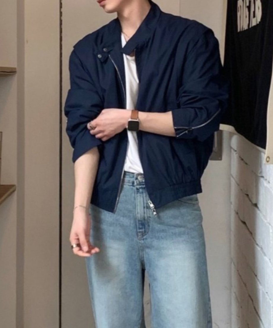 ネイビーのミニマルブルゾンにブリーチワイドデニムを合わせた男性の立ち姿。袖を引き上げて袖口のメタルジップディテールとバルーン調の袖のボリューム感が強調されたショット。