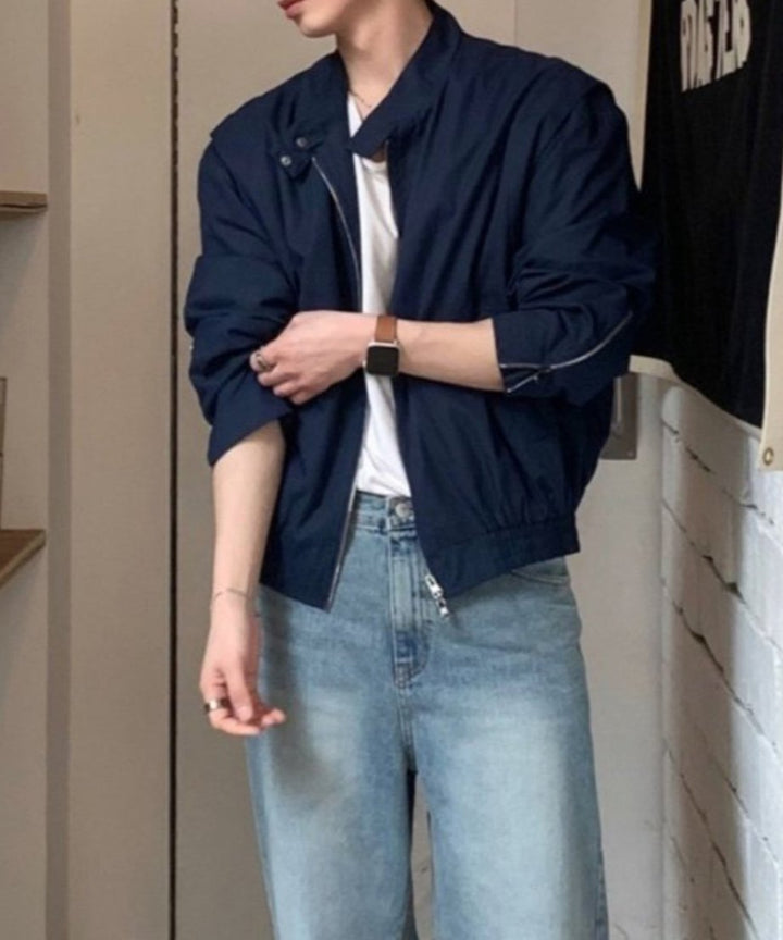 ネイビーのミニマルブルゾンにブリーチワイドデニムを合わせた男性の立ち姿。袖を引き上げて袖口のメタルジップディテールとバルーン調の袖のボリューム感が強調されたショット。