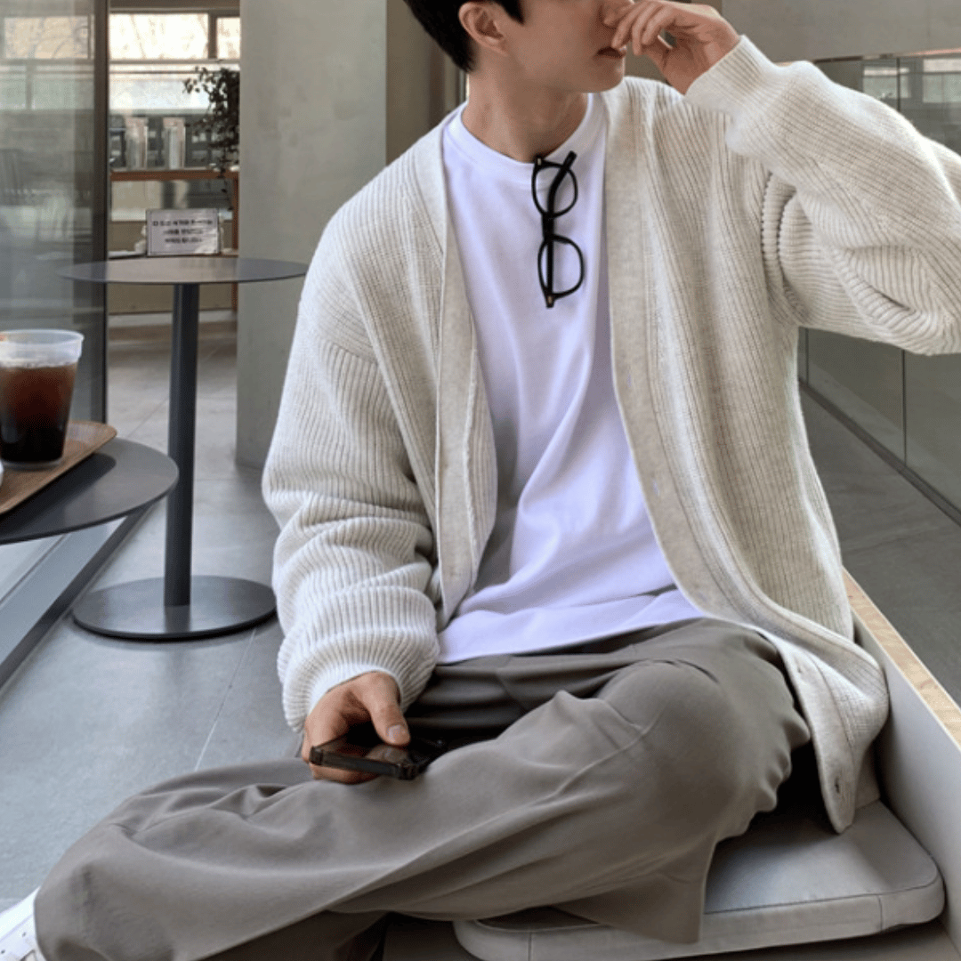 屋内のベンチシートに腰掛け、脚を組んでスマートフォンを右手で持ちながら、口元に手を当ててリラックスしている人物。ベージュのオーバーサイズカーディガンにニュアンスカラーのパンツを合わせた、クリーンな休日コーデ。