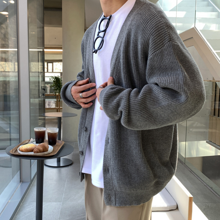グレーのオーバーサイズカーディガンを羽織った人物のサイドルック。両手を前立て部分に添え、ボリュームのある袖のたるみや、リブ編みの美しい立体感が際立つアングル。