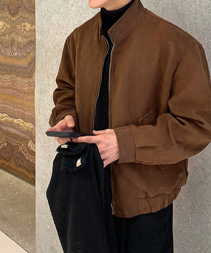 ブラウンのフェイクスエードブルゾンに黒のタートルネックを合わせ、片手にスマートフォンと黒のマフラーを持った男性のスタイリッシュな斜め前からのクローズアップカット。