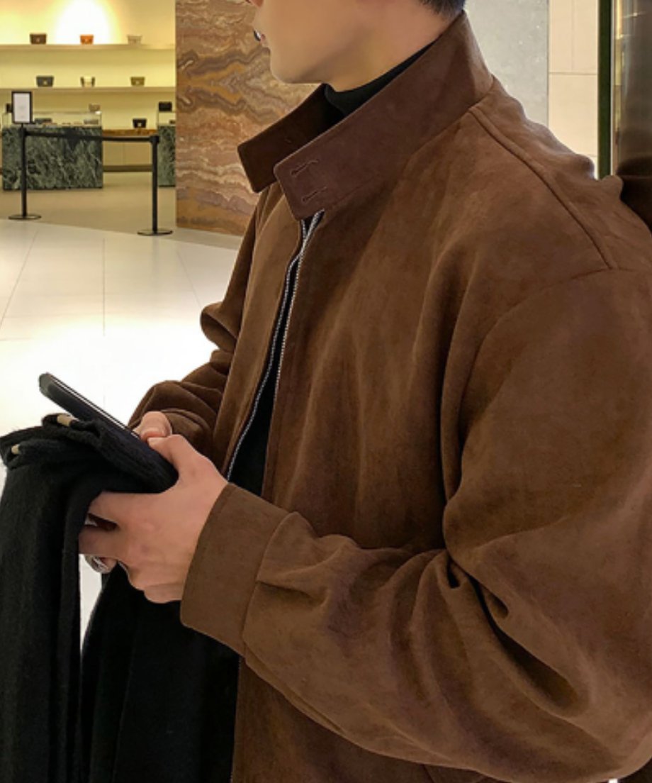 ブティックのような洗練された空間で、ブラウンのブルゾンを着用しスマートフォンを操作する男性のサイドからのクローズアップショット。スタンドカラーの美しい立ち上がりが特徴。