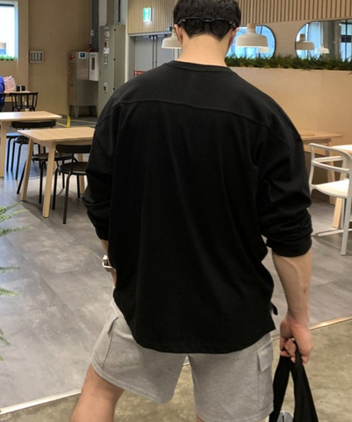 ブラックのオーバーサイズロンTを着用した男性の後ろ姿（バックスタイル）。頭の後ろに黒いサングラスを掛け、背中を横切るヨークシームデザインと、グレーのスウェットカーゴショーツを合わせた、開放的でモダンなストリート風のカフェ内カット。