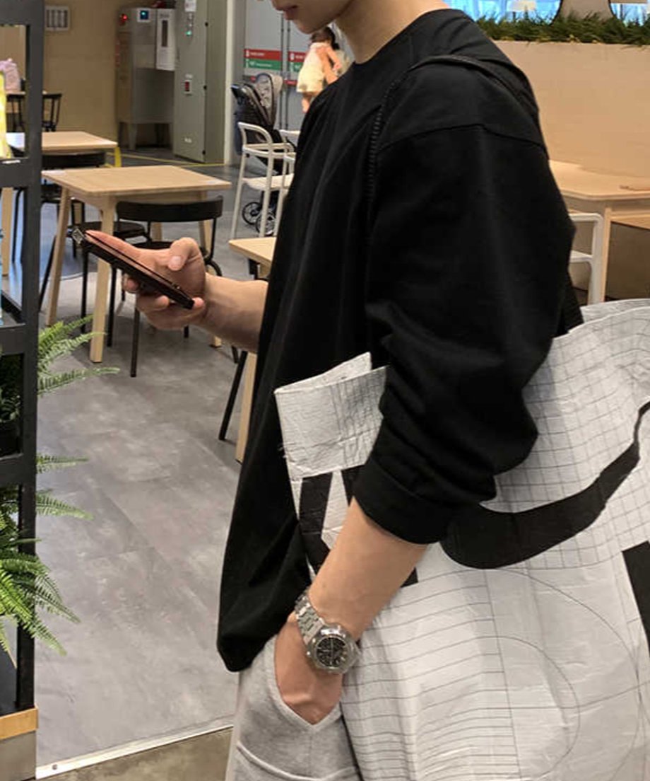スマートフォンを操作する手元にはメタルバンドの腕時計が輝き、白地に黒グラフィックの入った大容量トートバッグを肩掛けした、サイドスタイル。こなれ感のあるシティルック。