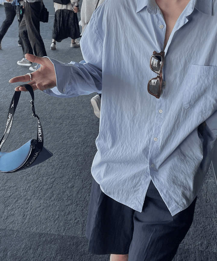 ライトブルーのシャツを着用した男性が、ロゴストラップ付きのブルーのミニポーチを指先に引っ掛けて見せる手元のパーツショット。ワッシャー加工シャツとショーツのネイビーのコントラスト。