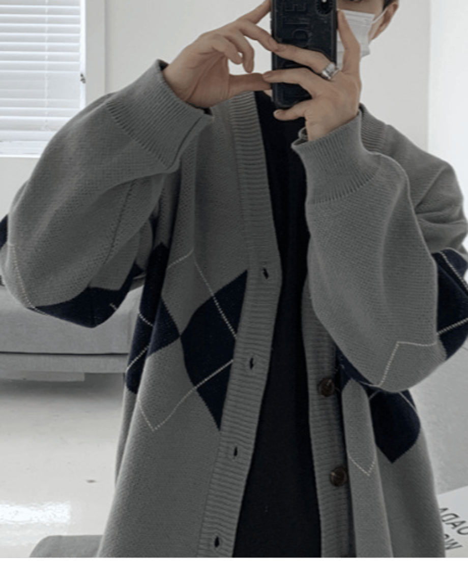 グレーのカーディガンを着用した男性が、スマートフォンを鏡に向けて自撮り（セルフィー）しているカット。太めの袖リブが手元にボリュームを作り、人差し指のシルバーリングが際立つスタイル。