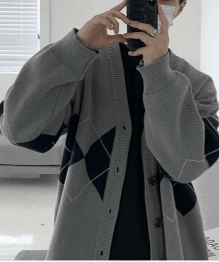 グレーのカーディガンを着用した男性が、スマートフォンを鏡に向けて自撮り（セルフィー）しているカット。太めの袖リブが手元にボリュームを作り、人差し指のシルバーリングが際立つスタイル。
