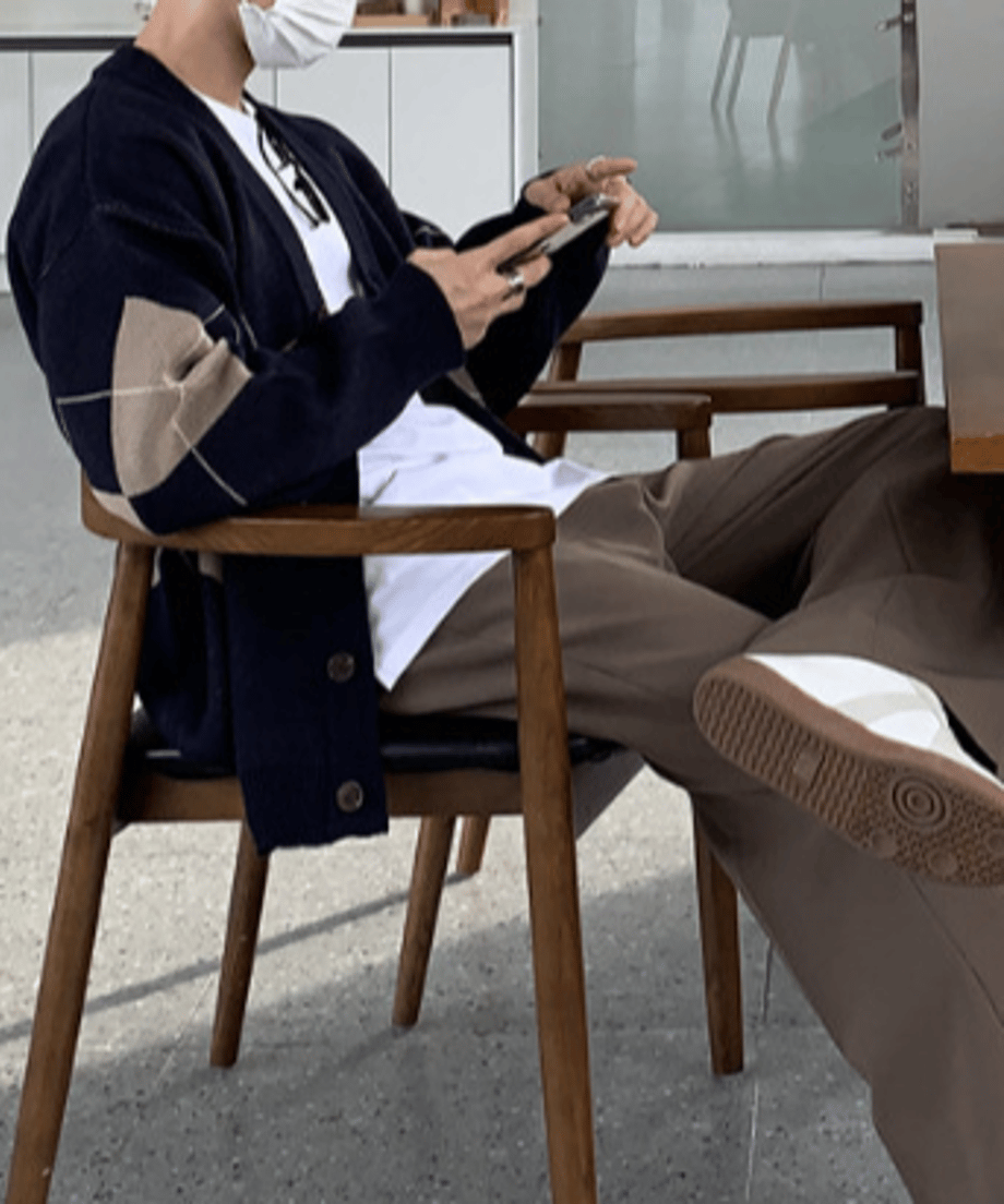 白いマスクをした男性が椅子に座り、スマートフォンを触る横顔に近いカット。ドロップショルダーによる袖のボリューム感と、ウッド調のインテリアがマッチしたリラックスルック。