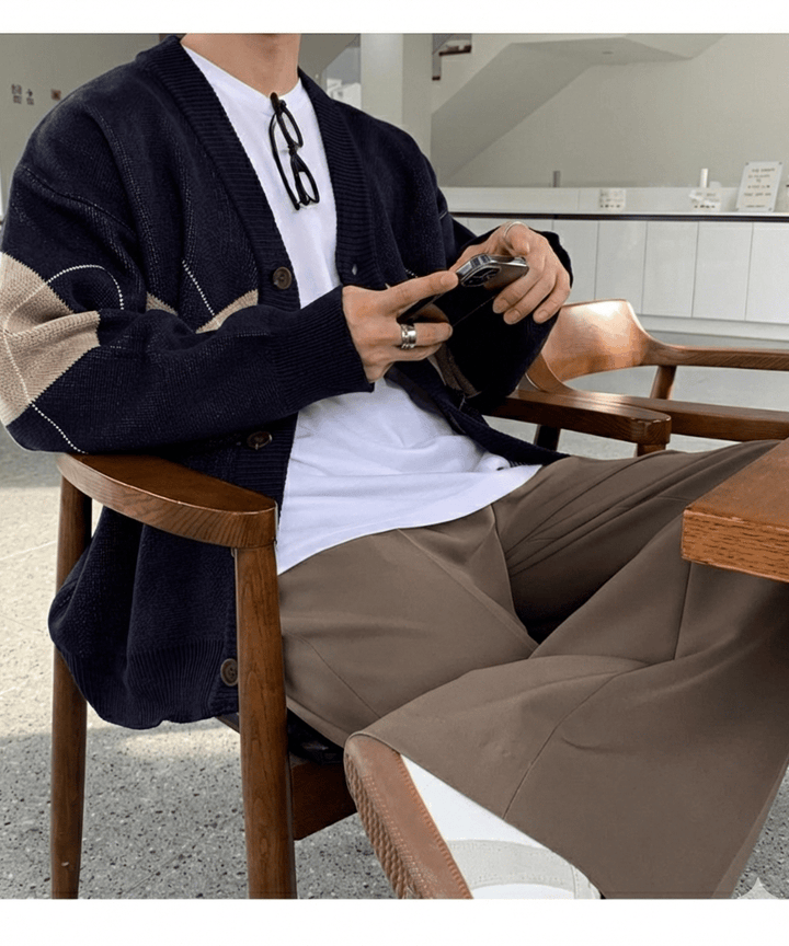 ネイビーのカーディガンに白いTシャツ、ブラウンのワイドパンツを合わせた男性が木製チェアーに深く腰掛け、手元でスマートフォンを操作しているミドルショット。胸元に掛けた黒縁メガネとシルバーリングがアクセント。