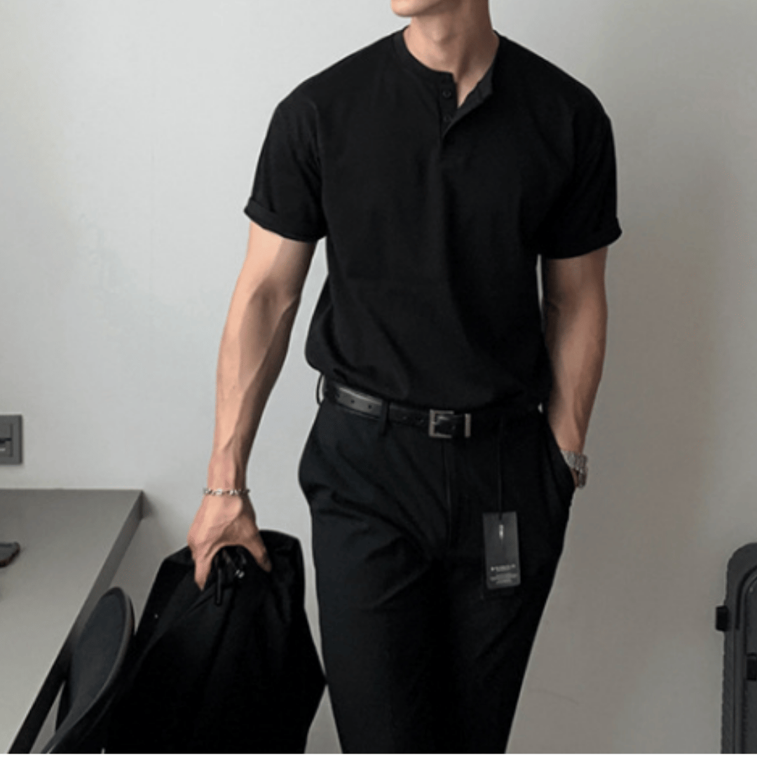 ブラックの半袖ヘンリーネックニットTシャツを黒のパンツにタックインし、左手をポケットに入れながら右手に黒のジャケットを持って立つ人物のフロントカット。