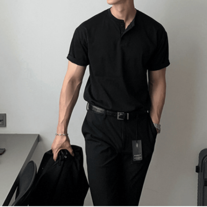 ブラックの半袖ヘンリーネックニットTシャツを黒のパンツにタックインし、左手をポケットに入れながら右手に黒のジャケットを持って立つ人物のフロントカット。