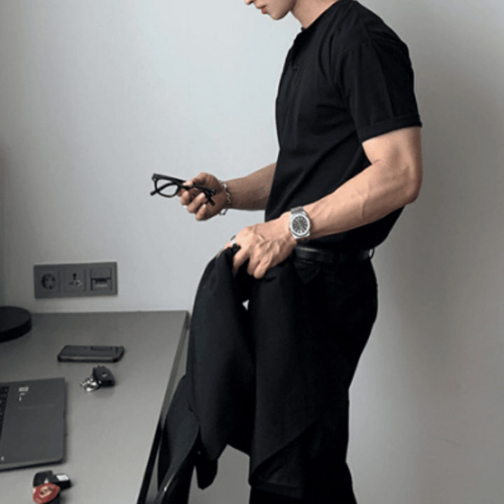 ブラックのヘンリーネックニットTシャツを着用した人物が、メタルバンドの腕時計を覗かせながら、右手にメガネと黒のジャケットを持ってデスクの横に佇む横顔のカット。