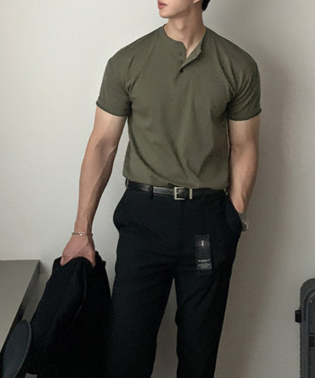 アーミーグリーンの半袖ヘンリーネックニットTシャツを黒のスラックスにタックインし、黒のレザーベルトを締めてジャケットを手に持った人物のフロントアップ。