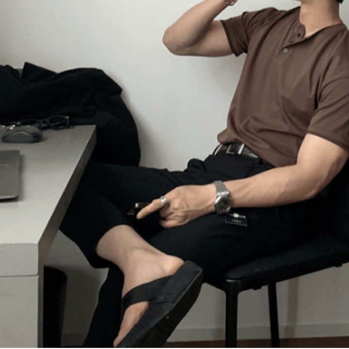 デスクの前に置かれた黒い椅子に斜めに腰掛け、脚を組んで右手で飲み物を飲むような仕草をする人物。コーヒーのヘンリーネックニットTシャツを黒のスラックスにタックインし、メタルバンドの腕時計と黒のサンダルを合わせたスタイリング。