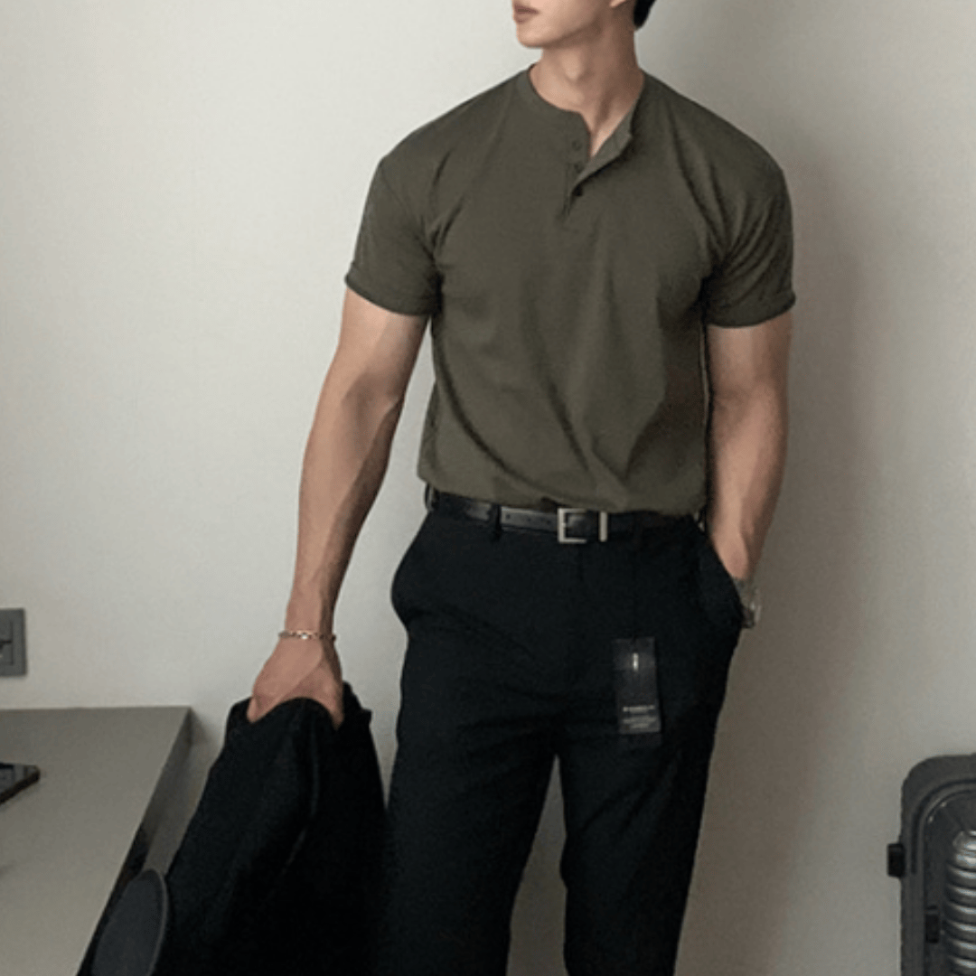アーミーグリーンの半袖ヘンリーネックニットTシャツを黒のスラックスにタックインし、黒のレザーベルトを締めてジャケットを手に持った人物のフロントアップ。