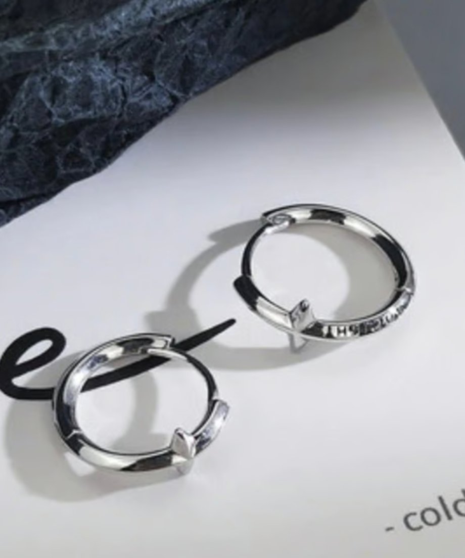 白い英字入りの紙の上に置かれた、輝くクロスモチーフ付きのシルバー製フープピアス。