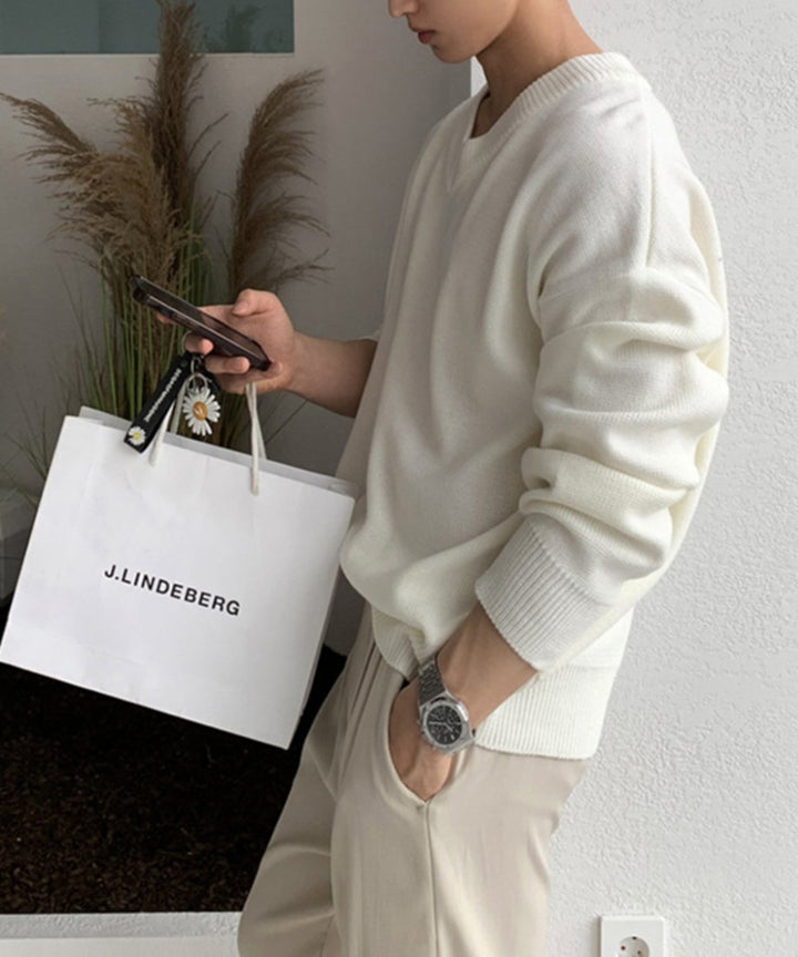 ホワイトのセーターにベージュのドレープパンツを合わせ、J.LINDEBERGの白いショップバッグとスマートフォンを手に持った男性のスタイリッシュな立ち姿。
