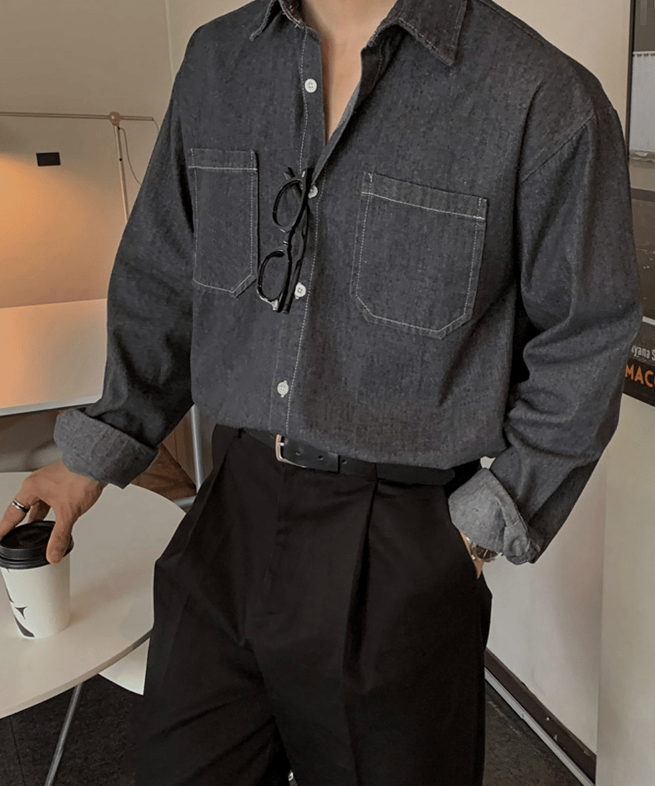 デニムステッチシャツを着用し、黒のベルト位置を両手で整える男性のチェストアップ。ドロップショルダーのゆったりとした袖を軽くロールアップした、こなれ感のあるディテールショット。