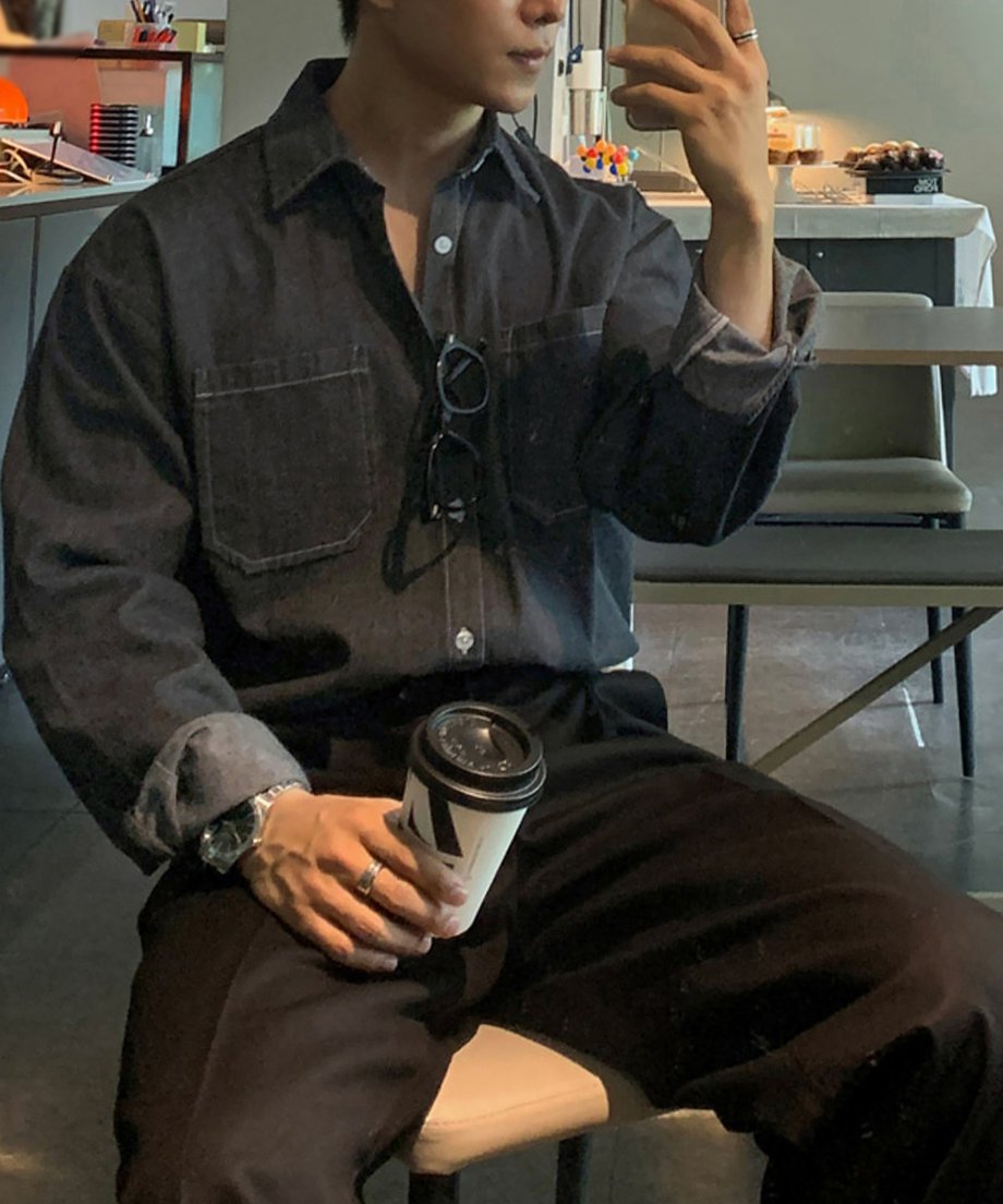 カフェの椅子に腰掛け、スマートフォンで鏡越しに自撮り（セルフィー）をする男性のスタイリングカット。ウォッシュドデニムシャツと黒のボトムスを合わせた、モダンなモノトーンの室内ルック。