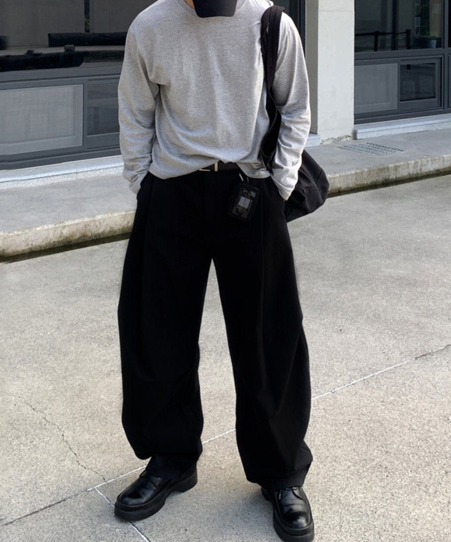 コンクリート調の屋外で、グレーのロンTをタックインし、大容量の黒バッグを肩掛けした男性の立ち姿。パンツの圧倒的なボリューム感と、全体の洗練されたストリートバランス。