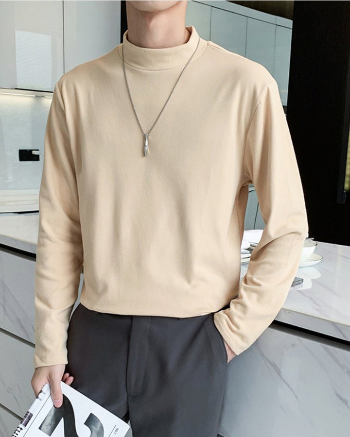 アプリコットのモックネック長袖Tシャツを着用し、カウンターに寄りかかる人物。チャコールグレーのスラックスを合わせ、右手には雑誌を持ち、首元にはシルバーのロングネックレスを着用。