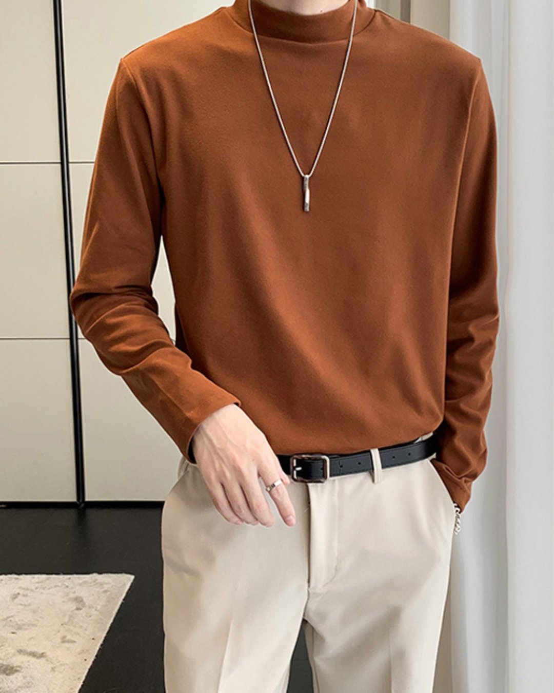 室内でコーヒーのモックネック長袖Tシャツを着用した人物のフロントカット。白のきれいめなスラックスに黒のレザーベルトを合わせ、首元にはシルバーのロングペンダントが掛けられている。