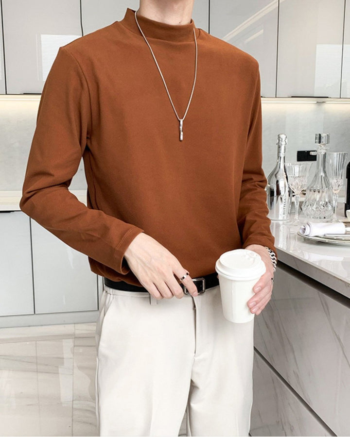深みのあるコーヒーのモックネック長袖Tシャツを着用した人物のフロントアップ。アイボリーのスラックスに、黒のレザーベルトをきっちりと締め、シルバーのロングペンダントを合わせたスタイリッシュなカラーコーディネート。
