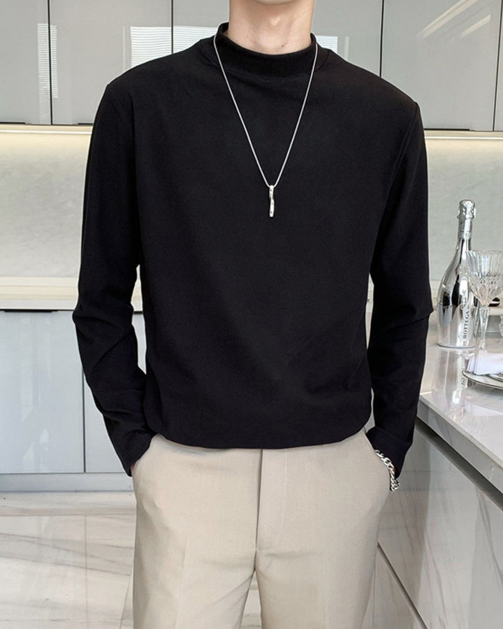 キッチンの前に立ち、両手をスラックスのポケットに入れたポーズの人物を捉えたカット。ブラックのモックネック長袖Tシャツとアイボリーのセンタープレスパンツ、シルバーのブレスレットを合わせた、スマートで都会的なコーディネート。