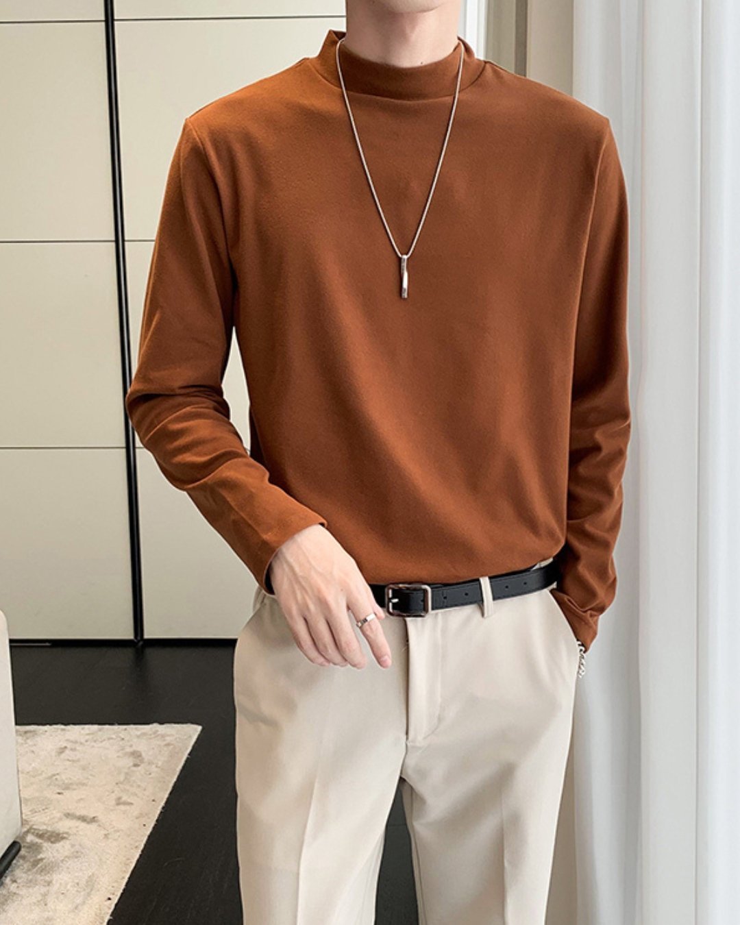 室内でコーヒーのモックネック長袖Tシャツを着用した人物のフロントカット。白のきれいめなスラックスに黒のレザーベルトを合わせ、首元にはシルバーのロングペンダントが掛けられている。