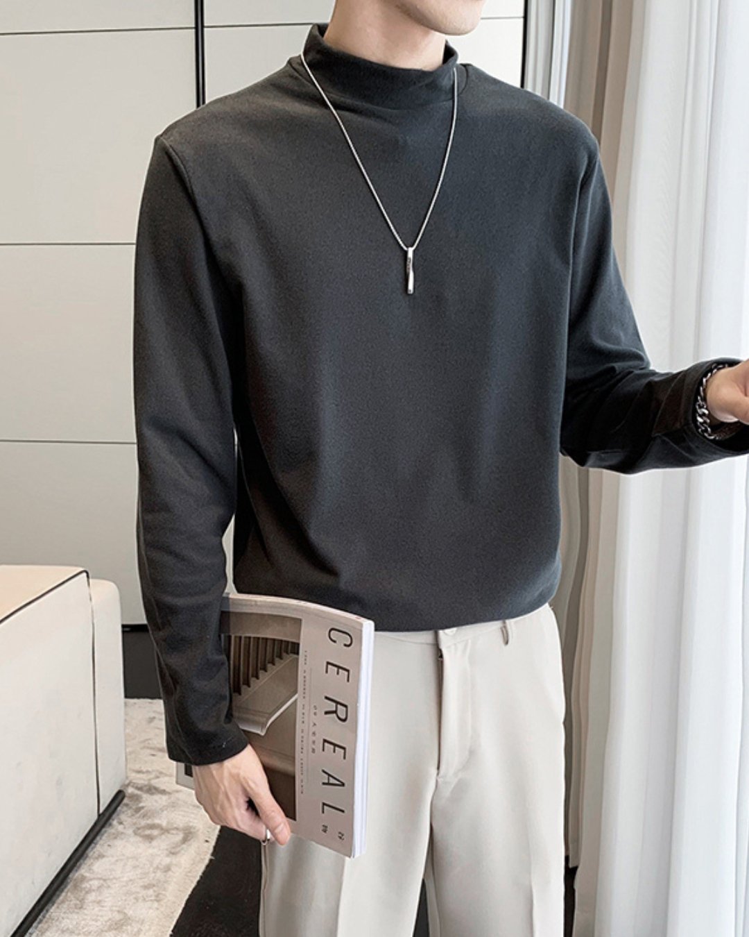 ダークグレーのモックネック長袖Tシャツを着用し、右手に雑誌を持つ人物のスタイリング。オフホワイトのスラックスにタックインした、上品で落ち着きのあるコーディネート。