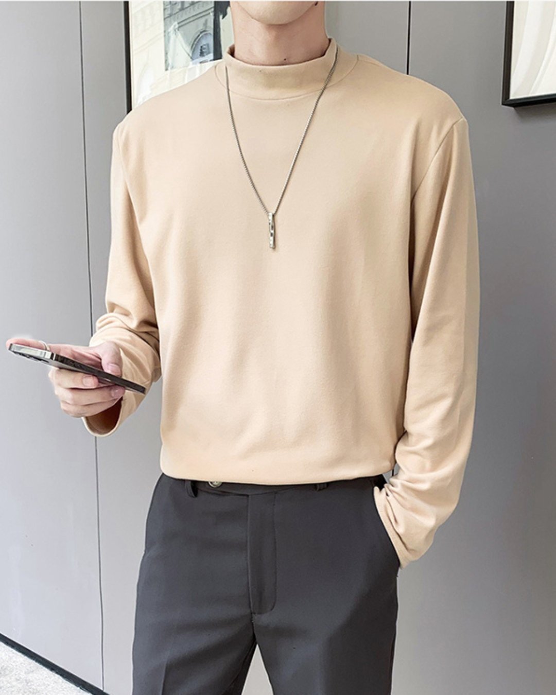 壁際に立ち、右手にスマートフォンを持つ人物のサイドルック。肌馴染みの良いアプリコットのモックネック長袖Tシャツを、チャコールグレーのスラックスにタックインした、洗練されたニュアンスカラーコーディネート。