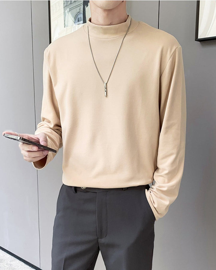 壁際に立ち、右手にスマートフォンを持つ人物のサイドルック。肌馴染みの良いアプリコットのモックネック長袖Tシャツを、チャコールグレーのスラックスにタックインした、洗練されたニュアンスカラーコーディネート。