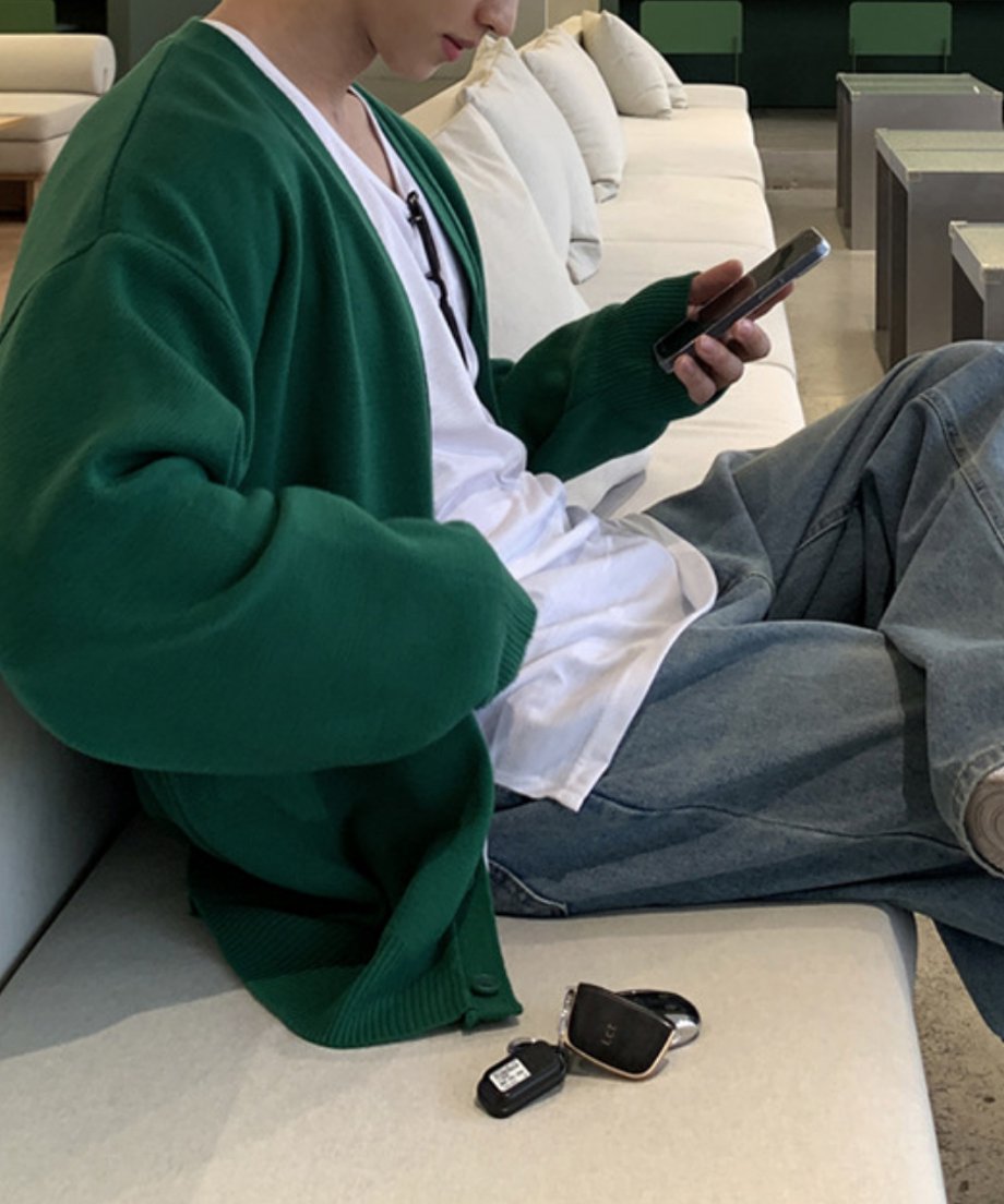 鮮やかなグリーンのプレーン編みルーズカーディガンに白Tシャツ、ルーズデニムを合わせ、カフェのソファ席でスマホを操作する男性のライフスタイルカット。手前には車のスマートキーが置かれている。