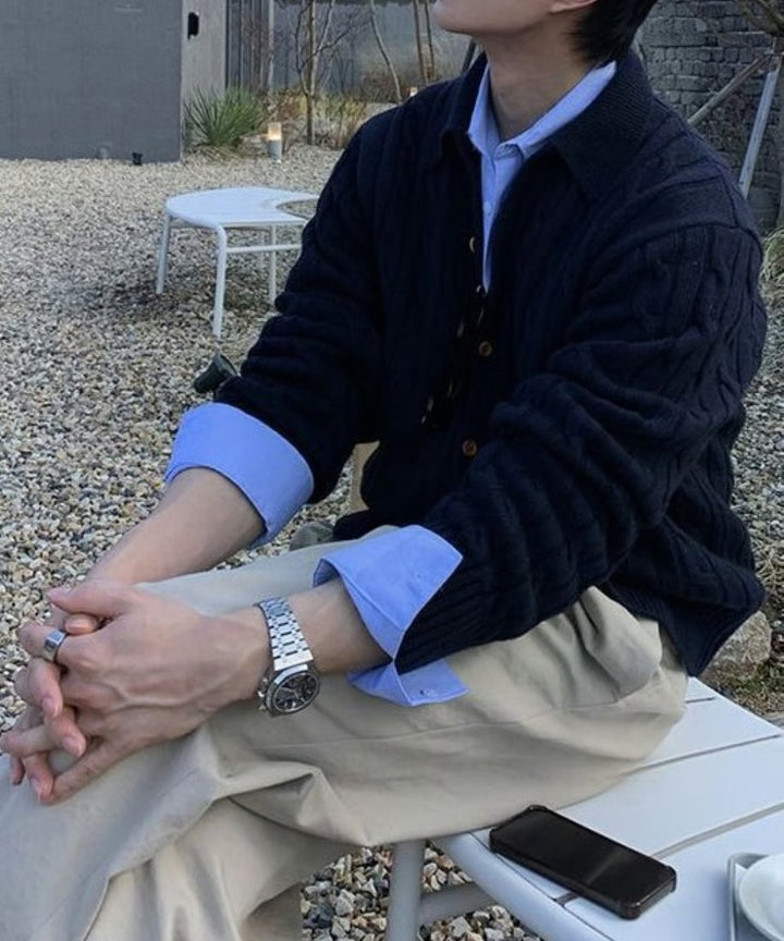 屋外の白いベンチに座り、手を組んでリラックスする男性のクローズアップカット。ネイビーのカーディガンとライトブルーのシャツ、ベージュのパンツのコントラストが美しい。