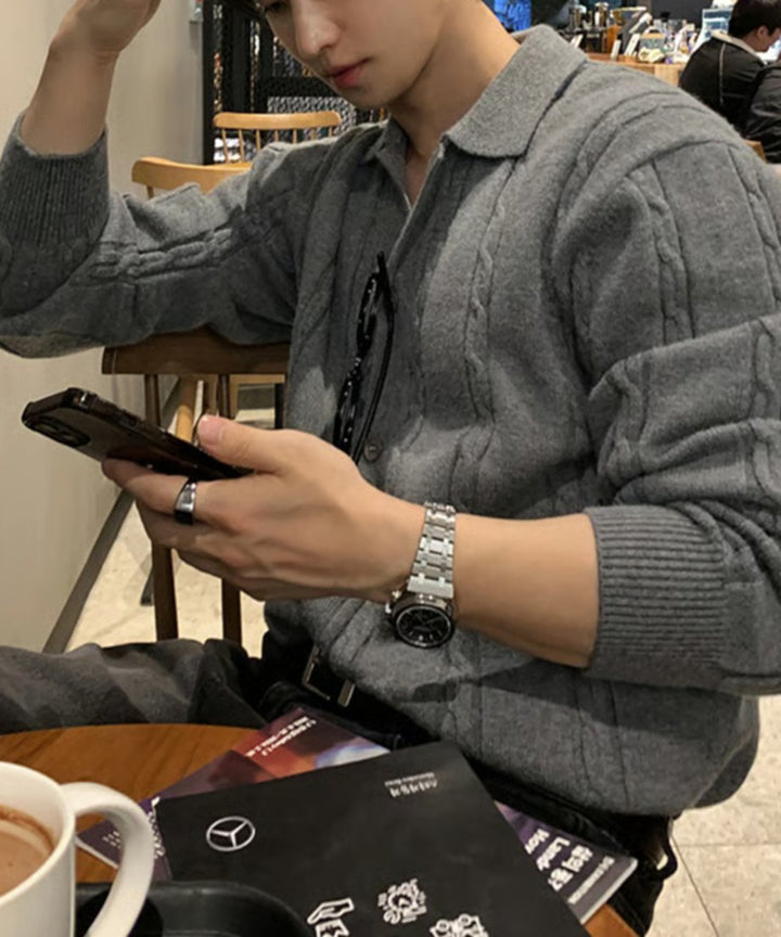 ダークグレーの襟付きケーブルニットポロカーディガンを着用し、カフェのテーブル席でスマートフォンを操作する男性のアップショット。手元にはシルバーの腕時計とリングが輝いている。