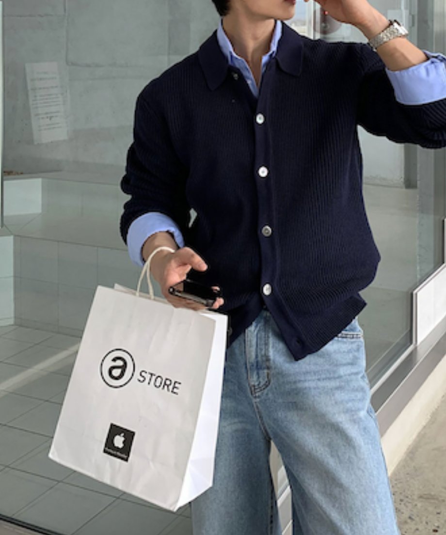 ネイビーのカーディガンを着用し、片手に「Apple Store」の白い紙袋、もう片方の手にスマートフォンを持った男性のフロントスタイリング。洗練された都会的なワンシーン。