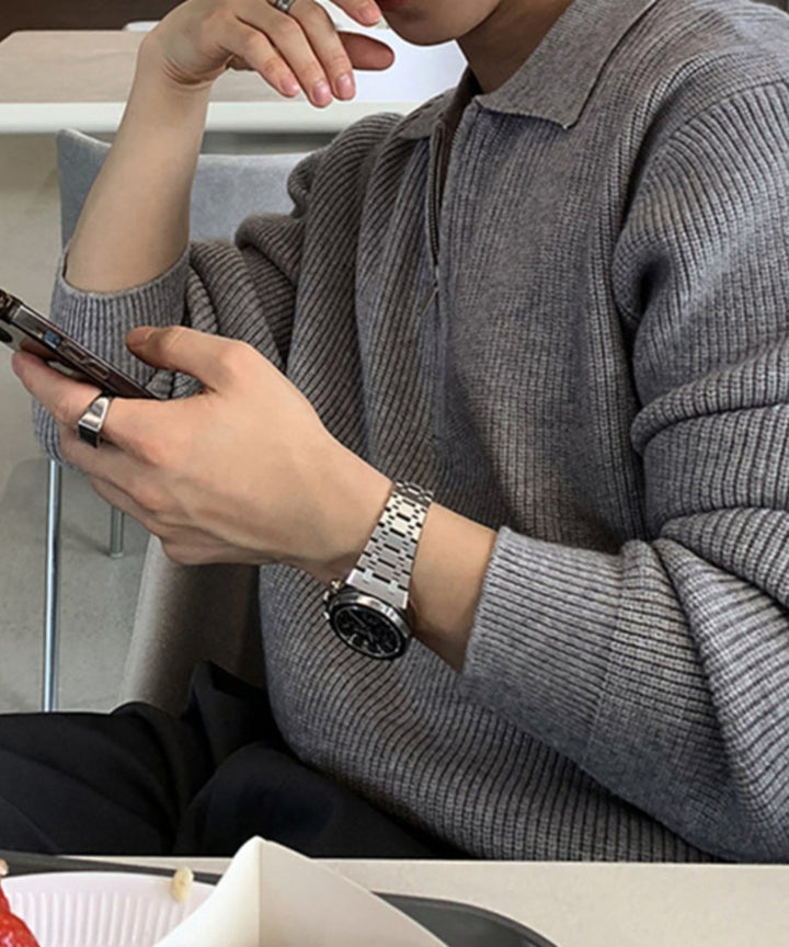 グレーのハーフジップリブニットを着用し、カフェの席でスマートフォンを操作する男性の手元と横顔のクローズアップ。メタルバンドの腕時計とシルバーリングが際立つ一枚。