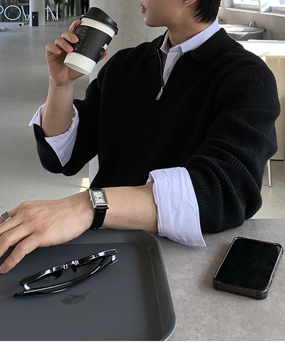 ブラックのハーフジップリブニットのインナーに白いシャツをレイヤードし、カフェのテーブルでコーヒーを飲む男性のミドルショット。手前には黒縁メガネとスマートフォン、黒いレザーストラップのスクエア腕時計が見える。
