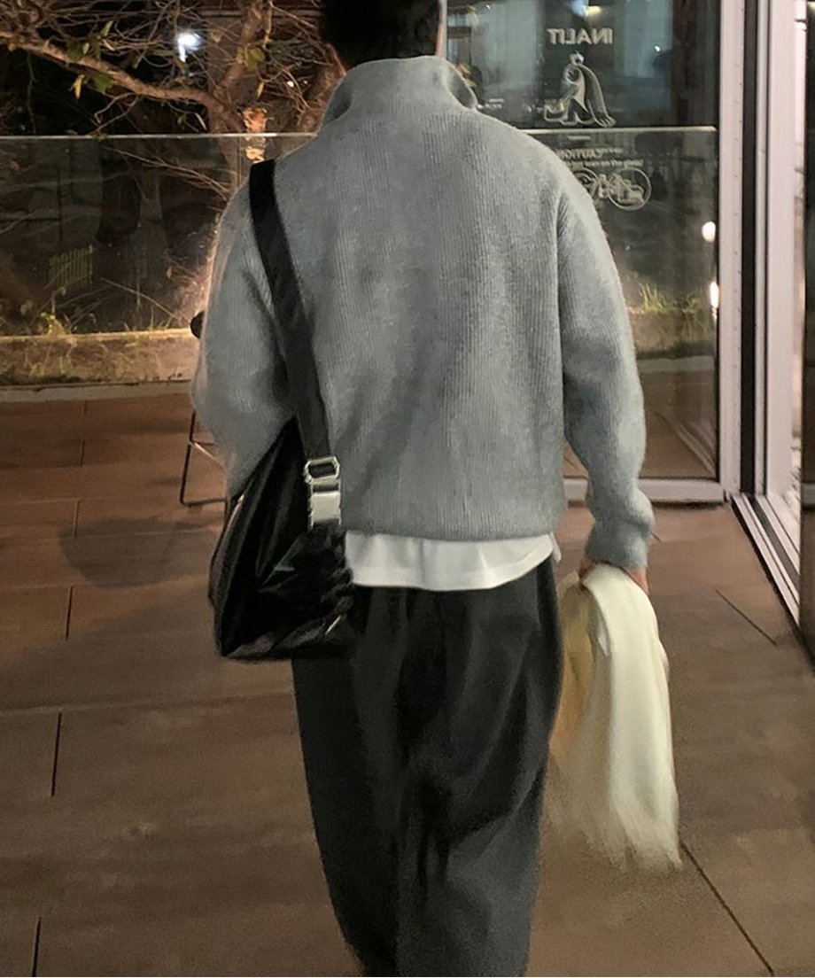 グレーのハーフジップニットに黒のレザーショルダーバッグを背負い、白いマフラーを手に持って歩き去る男性のバックスタイル。美しく並ぶ背中のリブ編みが確認できる。