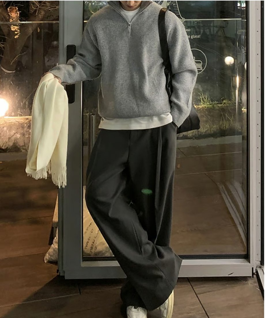 グレーのハーフジップニットにチャコールグレーのスラックス、スニーカーを合わせ、白いマフラーを手に持ってガラス扉の前に佇む男性の全身スタイリング。