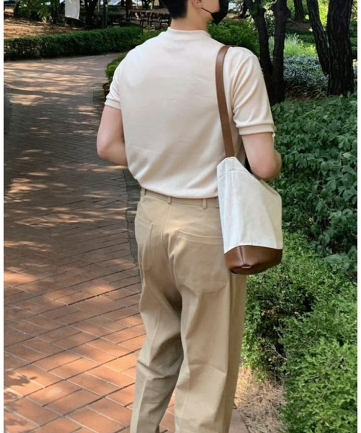 遊歩道を歩く男性のバック（背面）からのスタイリングカット。ニットポロのすっきりとした背面のシルエットと、肩から掛けたトートバッグ、ゆったりとしたベージュパンツの後ろ姿。