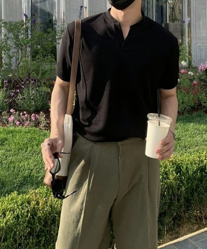 緑に囲まれた屋外で、サングラスを片手に持つ男性のスタイリングカット。ブラックのスキッパーニットポロにブラックのセンタープレスワイドパンツを合わせた、男らしく洗練されたカラーコーディネート。
