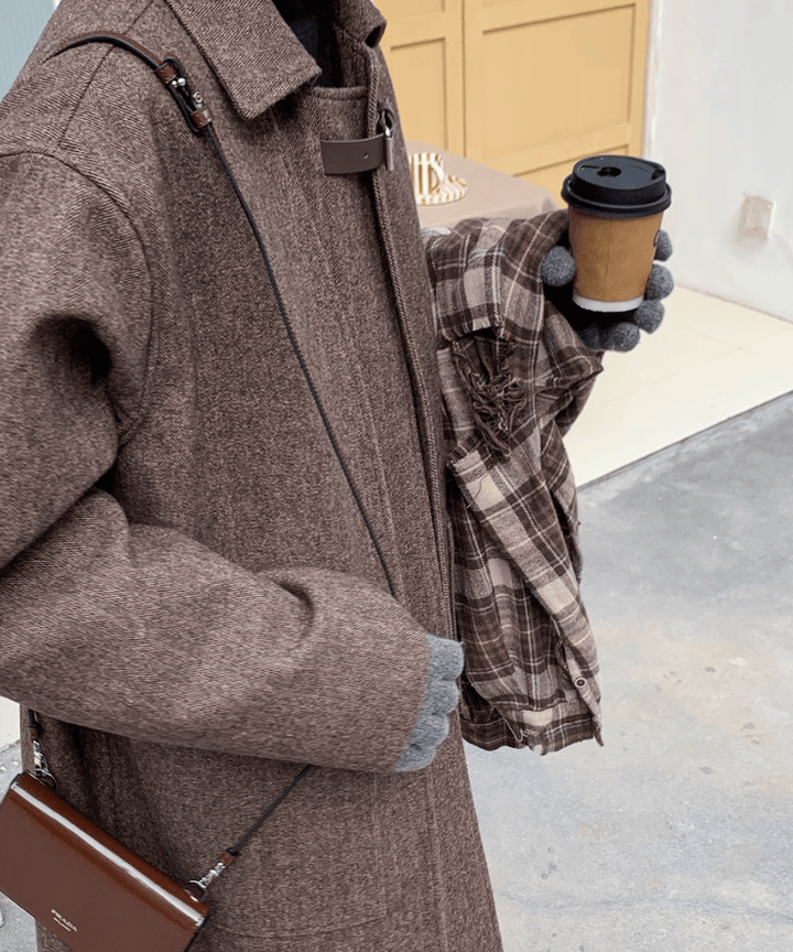 斜め前から撮影された、コートの生地とディテールのクローズアップ。メランジ感のある味わい深いウールブレンド生地の風合いや、袖口、ポケットの丁寧な仕立て、手に持ったコーヒーカップの質感がリアルに伝わるカット。